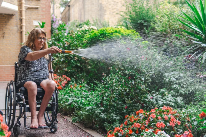 Credit: Centre for Ageing Better. A woman in a wheelchair sits by a blooming flowerbed, using a hose to water them.