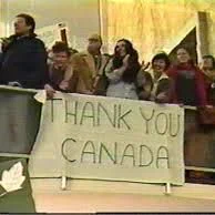A group of about 10-12 people standing on an upper balcony or mezzanine level indoors, leaning over a railing and waving enthusiastically toward the camera or below. Most are smiling or cheering, dressed in winter coats and casual clothing. In the foreground, they hold a large white banner with bold black handwritten text reading "THANK YOU CANADA" across two lines, with a small green maple leaf drawing on the left side. The background shows a beige or light-colored wall. The photo has a slightly grainy, older digital quality.