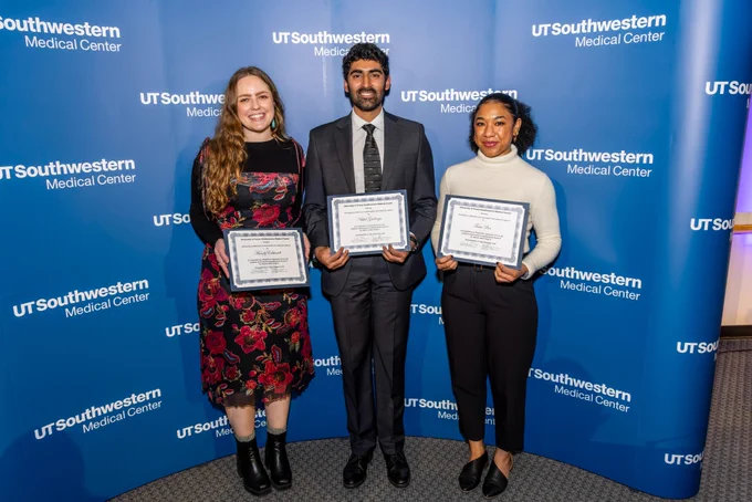 Three individuals holding certificates stand in front of a UT Southwestern Medical Center backdrop, all smiling. They are dressed formally for an award ceremony.