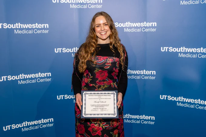 Person holding a certificate at UT Southwestern Medical Center event, smiling in front of a logo backdrop.