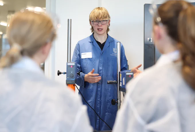 Members of the team from Unilever Port Sunlight instructing school children on how to conduct lab experiments. Unilever staff and students are all wearing lab coats and they are completing a formulation experiment on a large table with beaker, liquids and pipettes. 