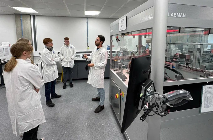 A team member from Unilever Port Sunlight instructs Year 12 students on how lab machinery works. The students are completing Unilever Port Sunlight's Year 12 Work Experience Programme. All staff and students are wearing lab coats and are standing in front of a large machine from 'LABMAN' - the machine is a large case with automated robotic arms inside.