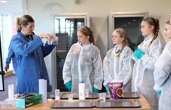 Members of the team from Unilever Port Sunlight instructing school children on how to conduct lab experiments. Unilever staff and students are all wearing lab coats and they are completing a formulation experiment on a large table with beaker, liquids and pipettes. 