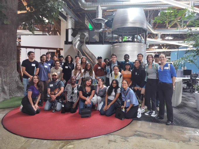 A group of people standing on a red carpet in an industrial-style office with a slide and an artificial tree.