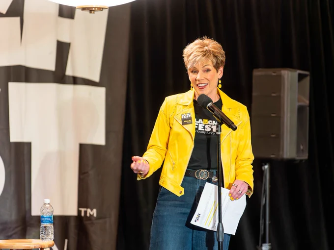 Person speaking into a microphone at a LaughFest event, wearing a bright yellow jacket with the festival's logo.