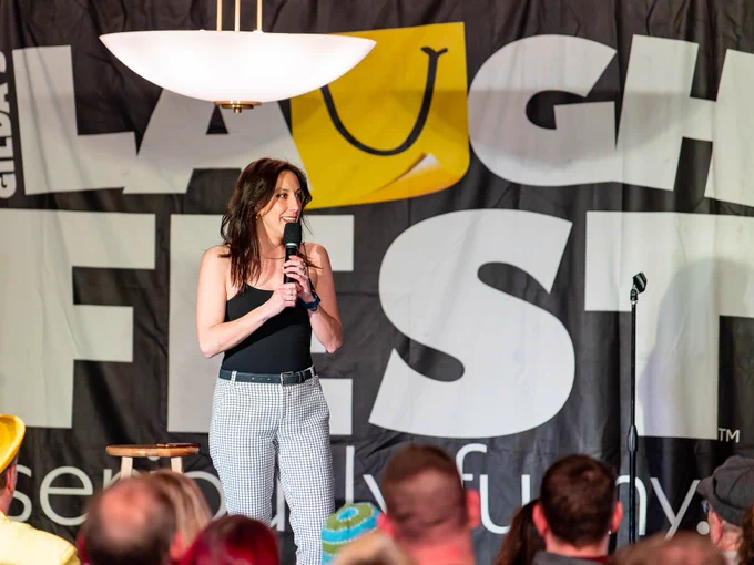 Person speaking into a microphone onstage at Gilda's LaughFest, facing an audience that includes someone wearing a yellow hat.
