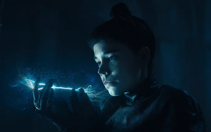 Laia Costa as Moghedien, her face lit by a cold blue glow as she studies a thin, glowing thread of the One Power in her hand, standing in a dark, shadowed space with an intense, calculating expression.