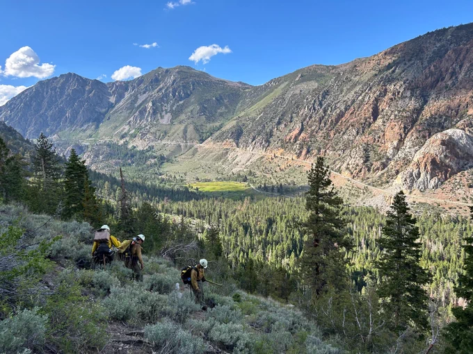 Three wildland firefighters wearing protective equipment hiking through a green mountainous landscape under a clear blue sky. Photo by Monte Kawahara. 