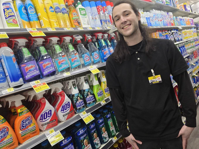 A store employee stands smiling in front of shelves stocked with cleaning supplies. He wears a black polo shirt. Rows of products such as Windex, 409, and Mr. Clean fill the shelves.
