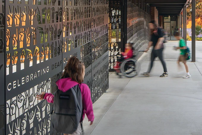 A person in a wheelchair and another with a backpack are passing by a decorative metal fence featuring various hand signs and the text "CELEBRATING DEAF CULTURE," alongside other pedestrians in motion. Photo by Lara Swimmer/Esto