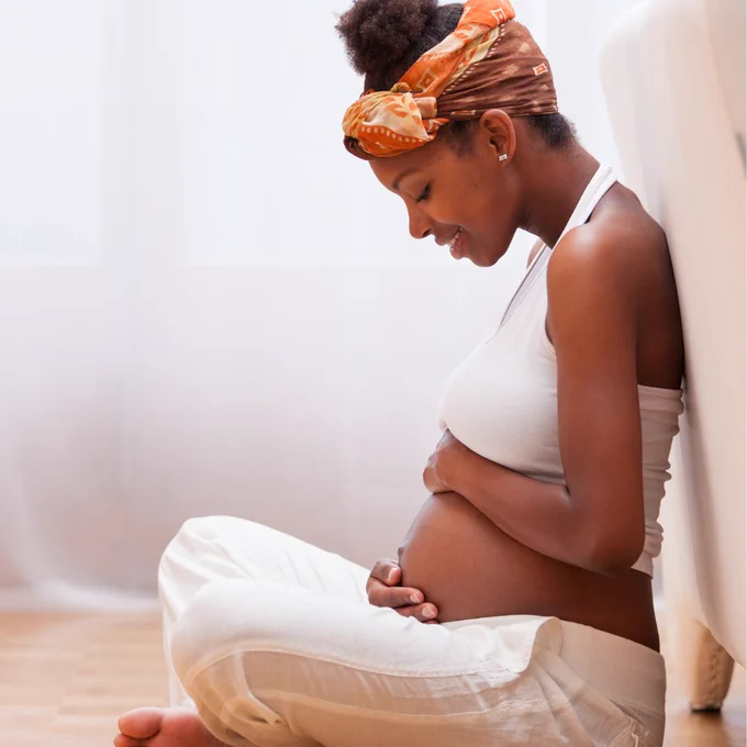 A woman sits on the floors, smiles, and holds her baby. 