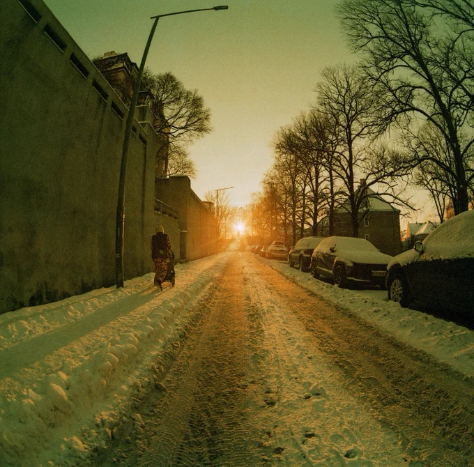 Sunset over a snow covered city street. My wife pushing a stroller on the pavement in a lot of snow, walking away from the camera. Snow covered cars on the other side.