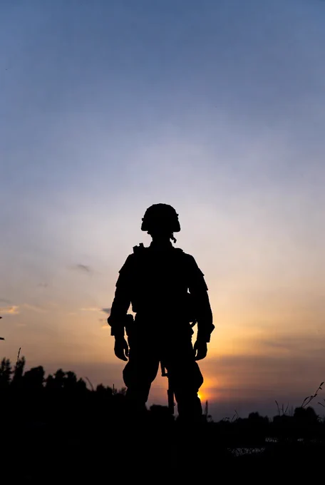 A quiet silhouette of a Ukrainian soldier standing still at dawn, seen from a distance. Soft blue morning light and warm tones on the horizon. Calm, realistic scene with no action, conveying readiness, resilience, and quiet connection.
