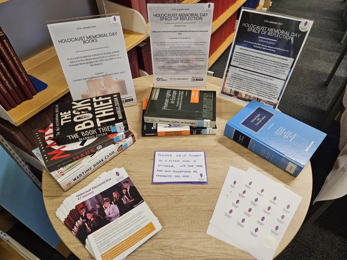A series of books relating to the Holocaust and human rights law are displayed on a round wooden table alongside resources from the Holocaust Memorial Day Trust. Posters featuring photos of white commemorative candles read 'Space of Reflection'. A handwritten note reads 'Please help yourself to a flyer and a sticker, we are here for any questions or thoughts you have'.