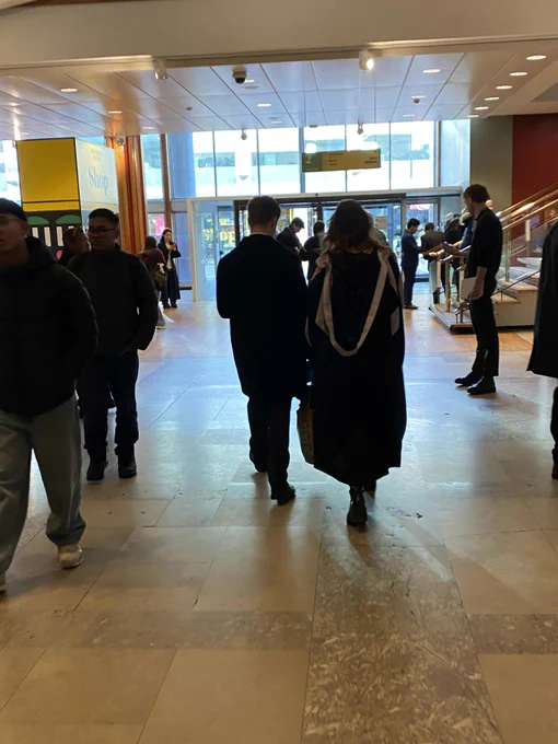 People in lavender trimmed graduation robes walk towards the hall, swooshing dramatically
