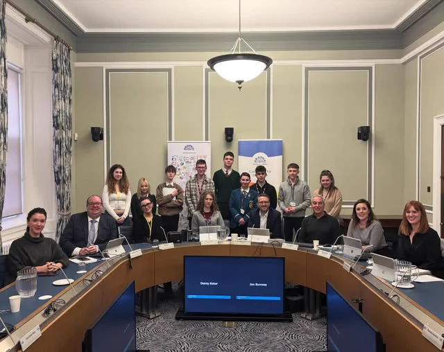 A group of young people gathered in committee meeting with MLAs. The room features green walls, white trim, and patterned curtains. The MLAs in attendance from left to right: Cara Hunter MLA, David Brooks MLA, Nick Mathison MLA (Committee Chair), Pat Sheehan MLA (Deputy Chair), Cathy Mason MLA and Michelle Guy MLA. A central screen on the table shows the names “Danny Baker” and “Jon Burrows.”