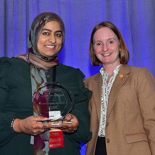 Two individuals smiling for the camera at an award ceremony, one holding a glass award, in front of a purple background.