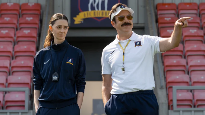  From Ted Lasso, Jason Sudeikis as Ted Lasso is pointing his left hand with Tanya Reynolds standing beside him on the soccer field.