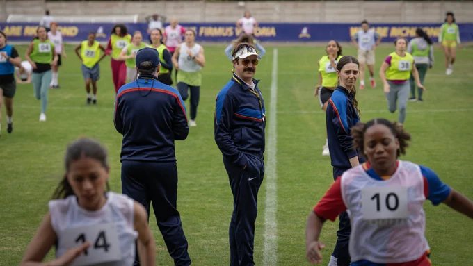 From Ted Lasso, Jason Sudeikis as Ted Lasso and Tanya Reynolds look back to the camera standing  on the soccer field with players practicing.