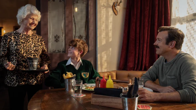 From Ted Lasso, Jason Sudeikis as Ted Lasso is seated with his son Grant Feely as Henry Lasso, while Annette Badland as Mae looks on smiling inside the pub.