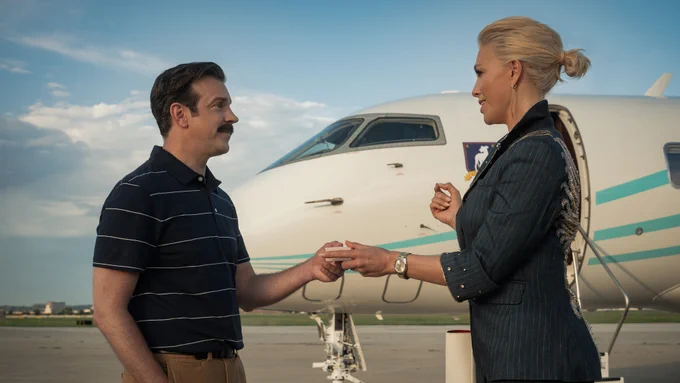 From Ted Lasso, Jason Sudeikis as Ted Lasso and Hannah Waddingham as Rebecca Welton exchange a box while on a runway in front of an airplane.