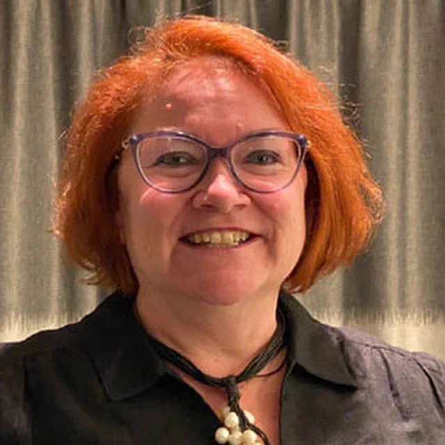 Ms Eisenhardt is smiling at the camera. She has red hair and is wearing glasses.