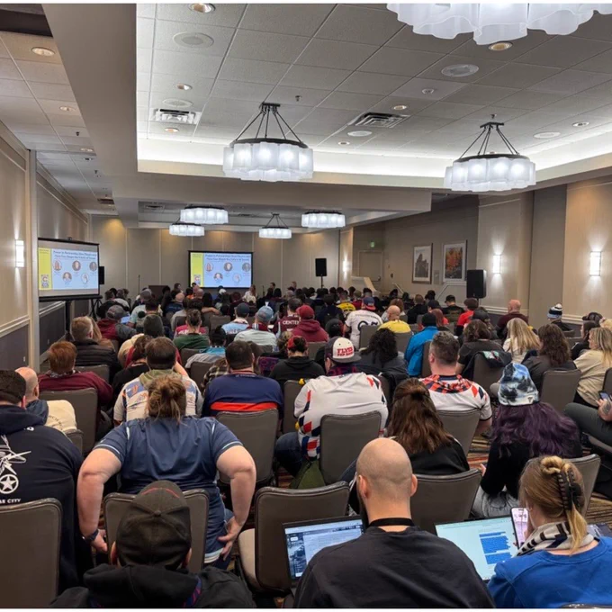 Large group of people seated in a conference room attentively watching a presentation on two screens at the front.