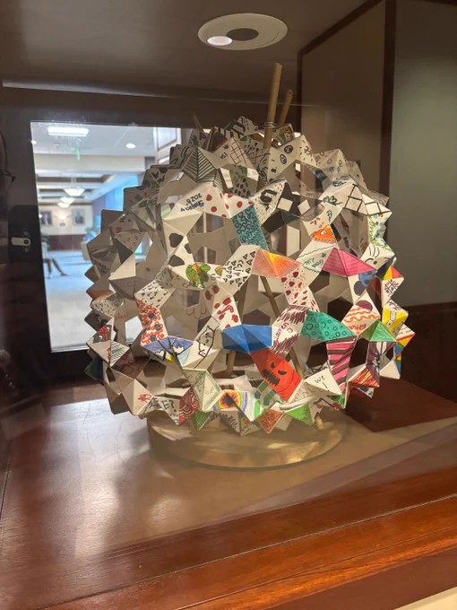 Intricate geometric paper sculpture with colorful patterns displayed on a wooden surface inside a glass case.