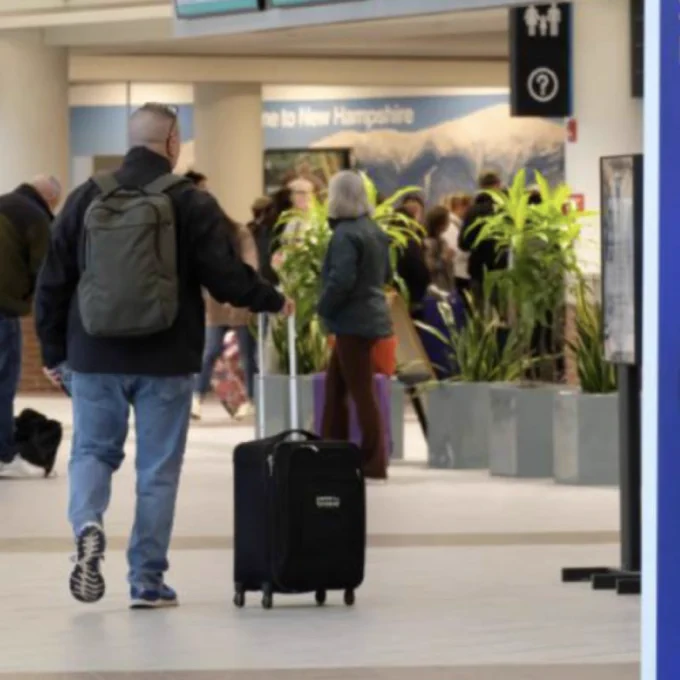A traveler with a backpack pulls a suitcase in an airport terminal. A self-service kiosk and a "Start Here" sign are in the foreground.