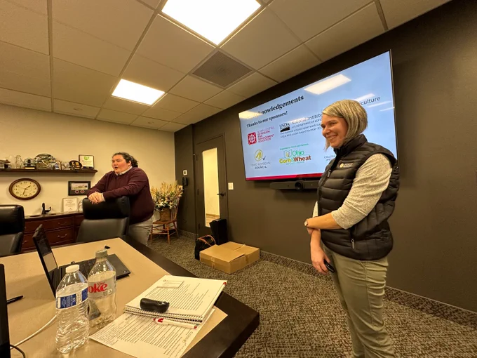 Photo of two women standing at the front of a conference room with a presentation on the tv behind them. The woman on the left has dark hair that is pulled back and is wearing a maroon sweater. The woman on the right has short grey hair and is wearing a grey shirt with a black vest.
