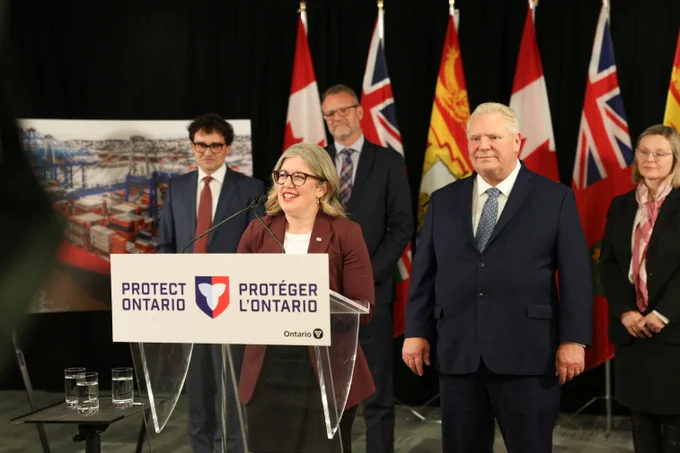 A woman speaks at a podium labeled "Protect Ontario" as several people, including a man in a suit, stand behind her. Multiple Canadian provincial flags are displayed in the background.
--
Une femme s'exprime sur un podium portant l'inscription "Protect Ontario", tandis que plusieurs personnes, dont un homme en costume, se tiennent derrière elle. Plusieurs drapeaux provinciaux canadiens sont déployés à l'arrière-plan.