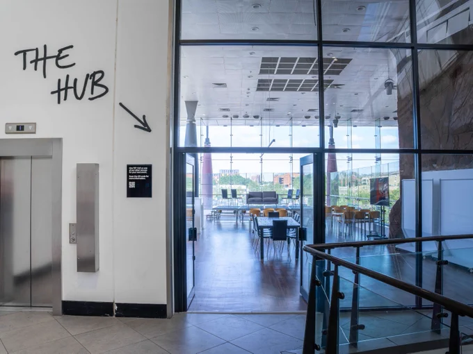 Interior of a spacious lobby featuring a sign that reads "THE HUB" with an arrow pointing to the right, a visible elevator on the left, and tables in a dining area through a glass wall at the back.