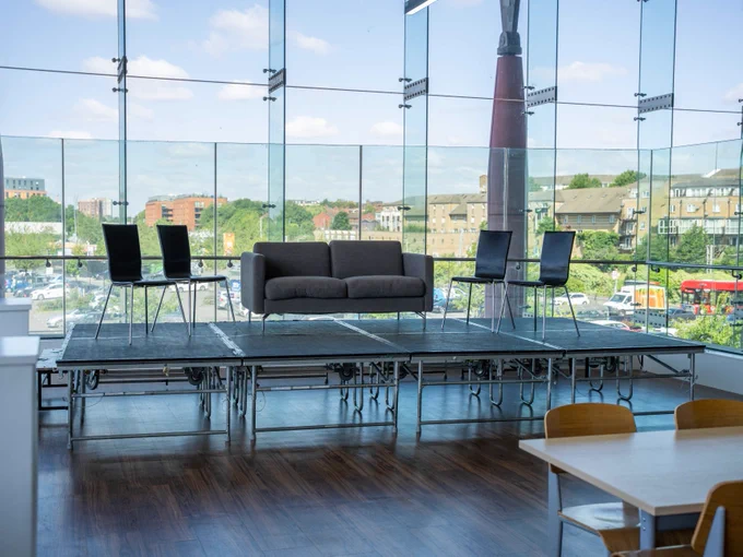 Modern office lounge area with a dark sofa and four chairs set against a backdrop of large windows showcasing a view of a cityscape. The space features a sleek, minimalist design with exposed ceiling pipes and natural lighting.