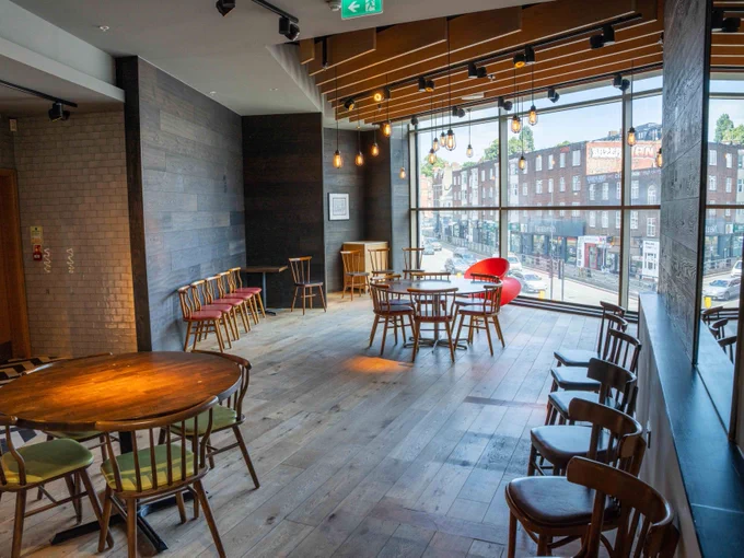 Interior of a spacious event space with various styles of chairs and tables, large windows overlooking a busy street, and modern lighting. Decor features a blend of wood floors, brick walls, and contemporary design elements.