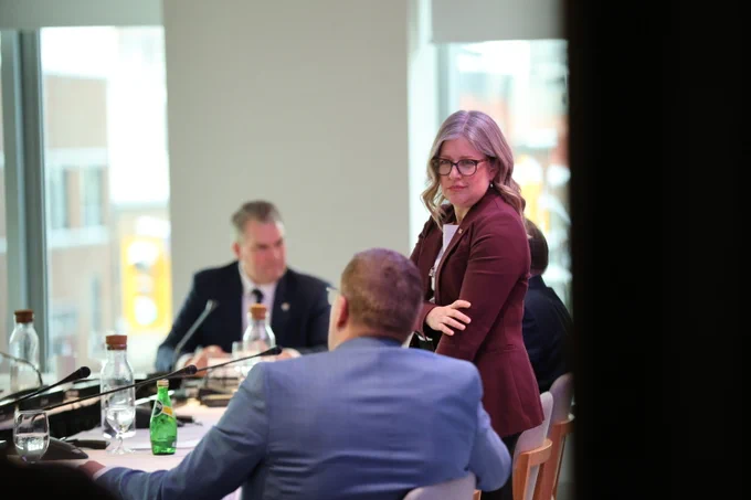 A woman in a maroon blazer stands and faces a man in a blue suit, who is seated at a conference table with microphones, bottles, and glasses. Another man is blurred in the background.
--
Une femme vêtue d'un blazer bordeaux se tient debout et fait face à un homme en costume bleu, assis à une table de conférence avec des microphones, des bouteilles et des verres. Un autre homme est flou à l'arrière-plan.