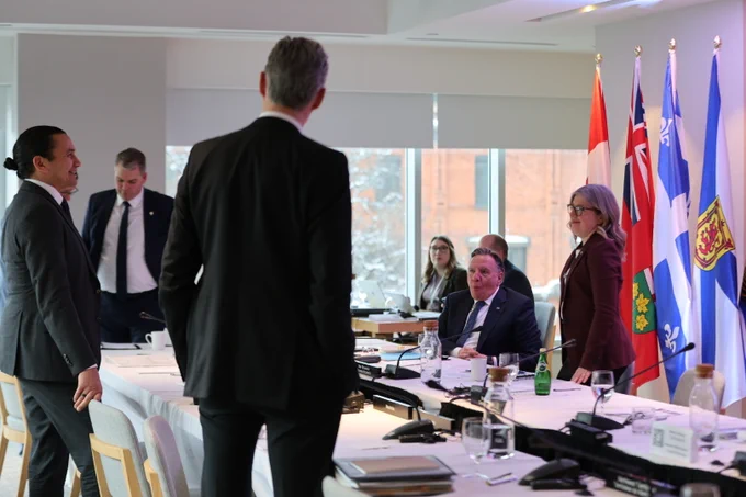 A group of people in business attire stand and sit around a conference table with documents and microphones. Five provincial flags are displayed in the background by large windows.
--
 Un groupe de personnes en tenue de travail se tient debout et assis autour d'une table de conférence avec des documents et des microphones. Cinq drapeaux provinciaux sont exposés à l'arrière-plan par de grandes fenêtres.