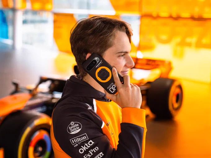 Oscar in profile inside the McLaren garage, wearing an orange and black team hoodie, holding a phone to his ear with a black and orange Quad Lock OP81 phone case visible. A McLaren car is blurred in the background.