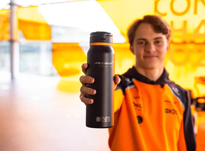 Oscar standing in the McLaren garage, wearing an orange team top and smiling slightly while holding a black McLaren MAG bottle with Quad Lock branding toward the camera, with the background softly out of focus.