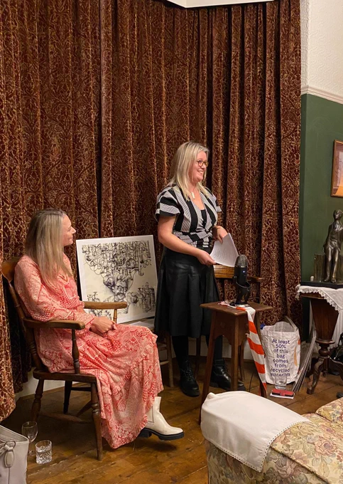 Two white women are in front of drawn red curtains. One is standing, the other is sat down. There is a wooden floor and a sofa.