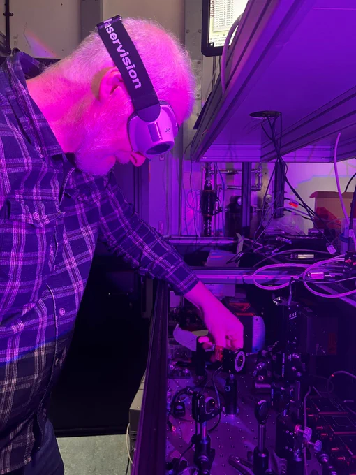 A scientist wearing goggles works on an optics table. the image is purple hued.