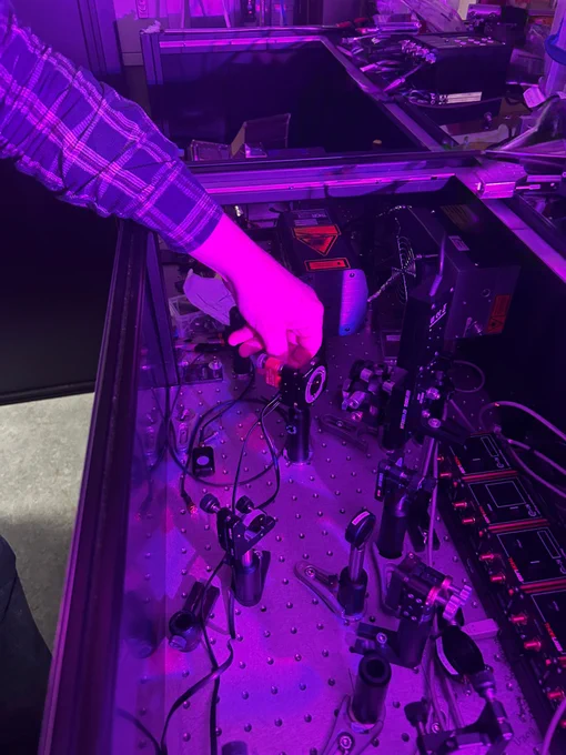 A scientist wearing goggles works on an optics table. the image is purple hued.