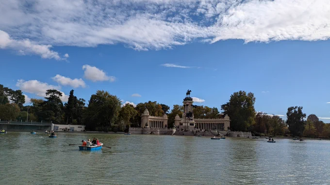 El estanque del Retiro, con barquitas navegando y el cielo azul con nubes blancas. Al fondo, árboles y el monumento a Alfonso XII.