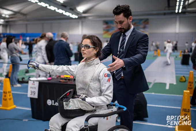 A dark-haired fencer in foil fencing kit sits in a wheelchair, arm outstretched and holding their mask. The referee stands behind them, measuring distance before the match begins. An electronic scoring system can be seen behind, as well as a competition environment with standing fencers competing and other spectators.