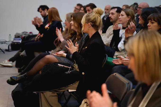 A number of people listening to a conference sitting on black metal chairs, all facing the same direction and clapping their hands.