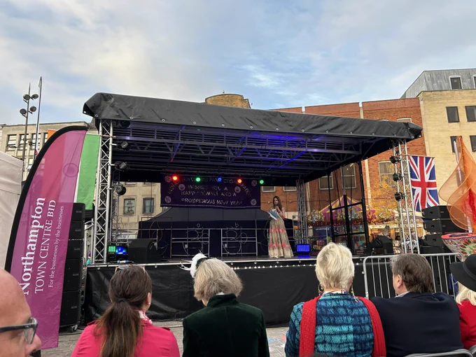 Image shows the stage at Northampton Diwali 2024. The stage backdrop is black with silver supporting posts. At the back of the stage a banner reads Indian Hindu Welfare Organisation wishes you a Happy Diwali and a Prosperous New Year