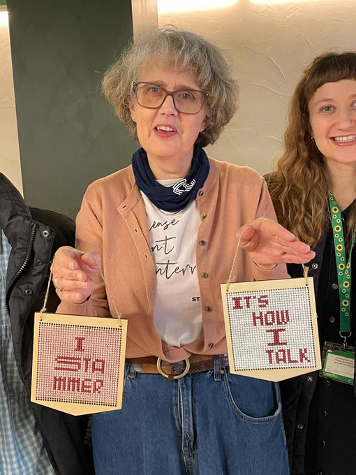 The photo is of 2 women who stammer. One is holding 2 wooden cross stitch pennants saying I stammer and It's How I Talk