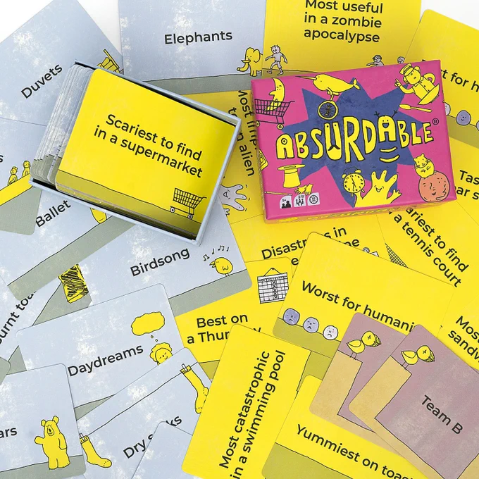 An open box of Absurdable, a card game, sitting on a table covered in cards. The box has a bright pink lid that says Absurdable in big, fun letters, and it has several funny illustrations on it. The cards are also illustrated and read things like: daydreams, birdsong, scariest to find in a supermarket, elephants, most useful in a zombie apocalypse, duvets, etc.