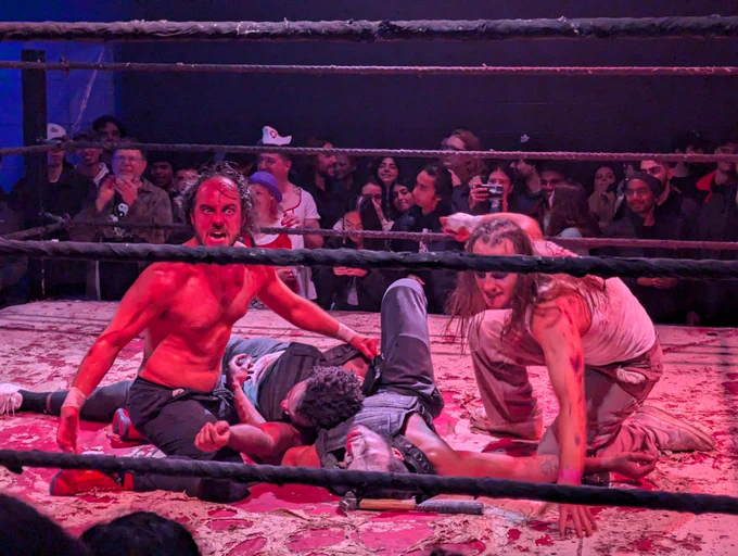 Two wrestlers dressed like zombies pose over two other wrestlers in a wrestling ring