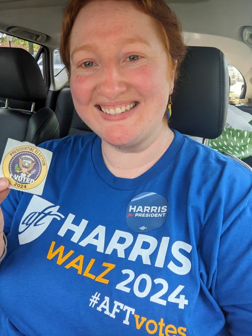 Me posing in my car with a sticker that reads "I Voted: presidential election 2024". I am wearing a blue shirt that reads "AFT Harris Walz 2024 #AFTvotes" and am wearing a "Harris for President" sticker.