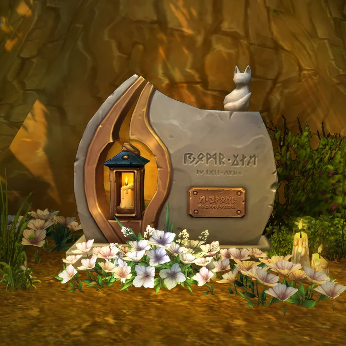 An image of the in-game memorial for Ibelin, located on an island in Crystal Lake in Elwynn Forest. The large stone tablet sits at the base of a large tree with white flowers growing around it. The tablet is rounded on each side, and extends higher on the left side, with an opening that houses a small lantern with a burning candle. Azerothian text is inscribed within the stone, with a copper plaque below and additional text. A stone statue of a small fox sits atop the right side.
